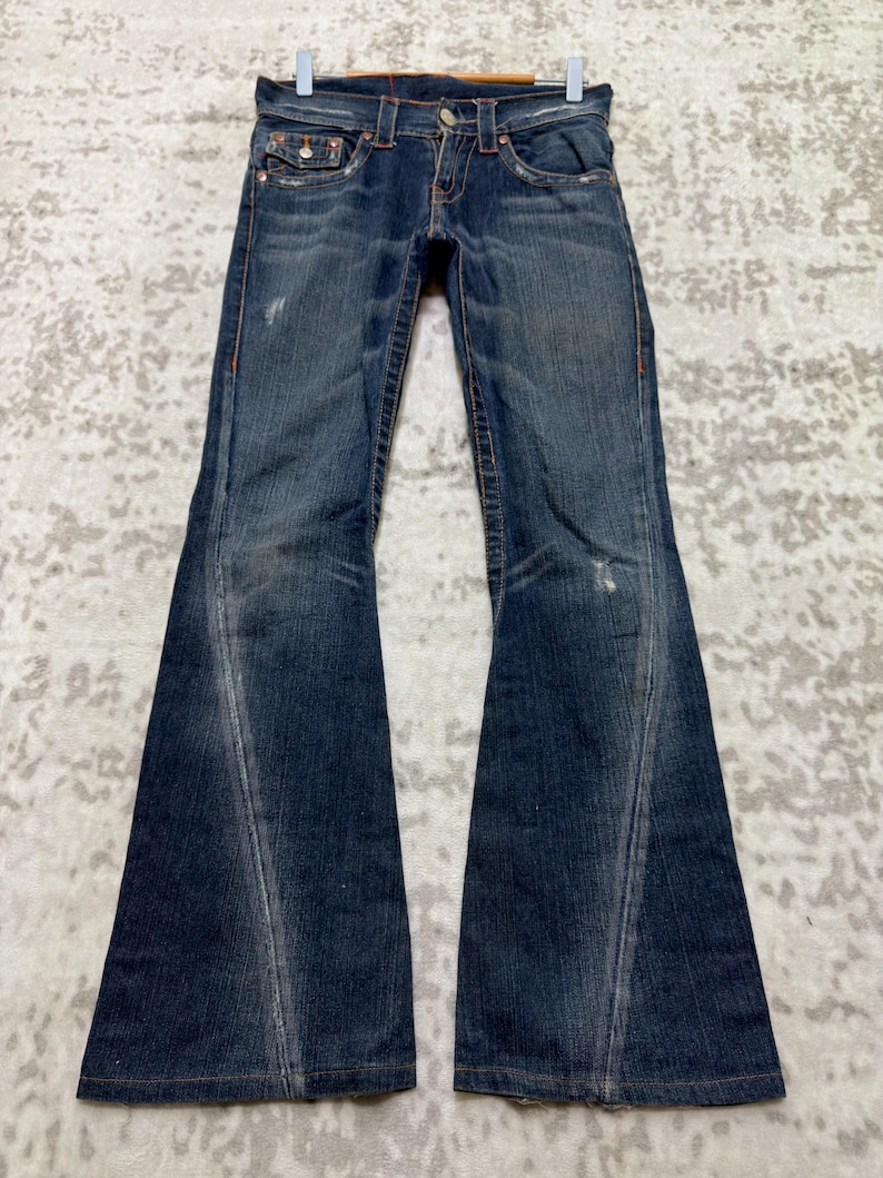 May include: Dark wash denim jeans with a flared leg design. The jeans feature a classic five-pocket style, red stitching, and a button and zipper closure. The jeans are hanging on a white hanger.