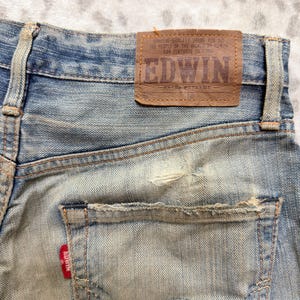 Puede incluir: Primer plano de vaqueros de mezclilla azul desgastados. Los vaqueros presentan un parche de cuero marr&oacute;n con la marca "EDWIN" y texto. Una etiqueta roja con el nombre de la marca es visible en el bolsillo trasero. Los vaqueros tienen un aspecto desgastado y descolorido.