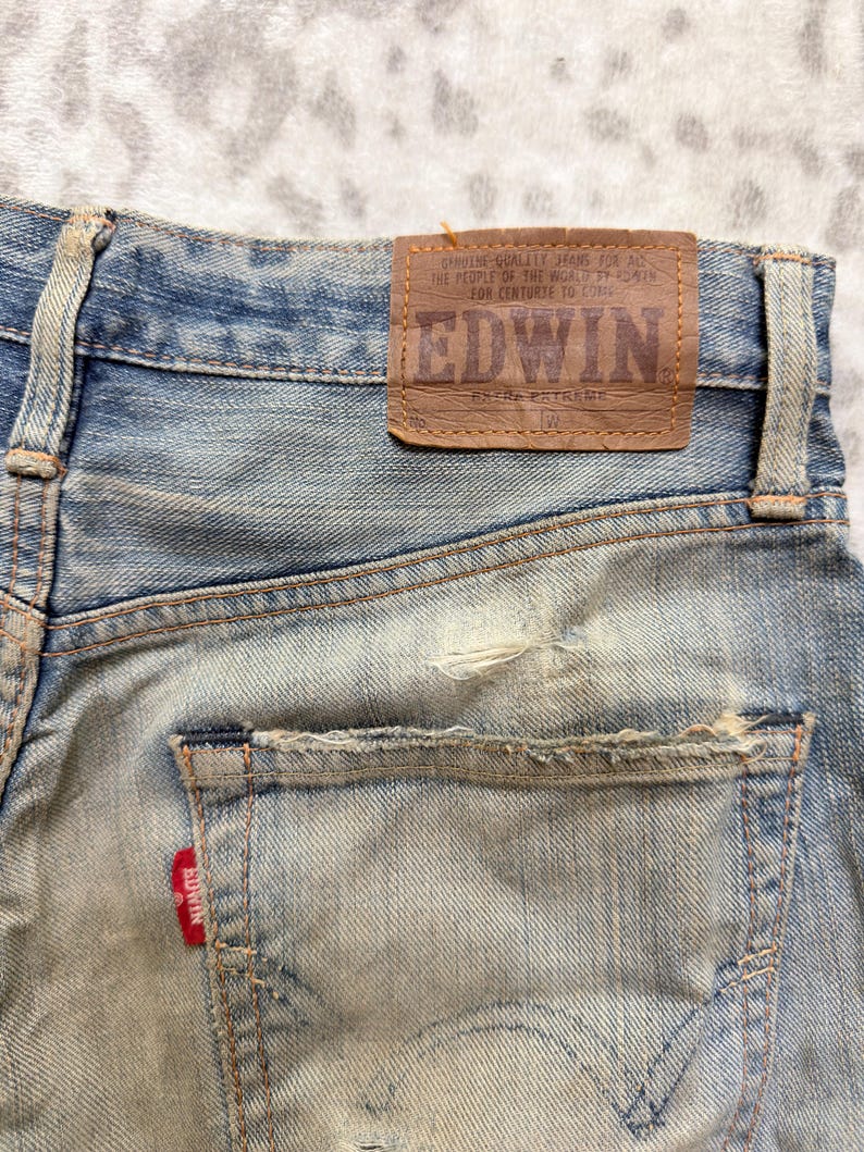 Puede incluir: Primer plano de vaqueros de mezclilla azul desgastados. Los vaqueros presentan un parche de cuero marr&oacute;n con la marca "EDWIN" y texto. Una etiqueta roja con el nombre de la marca es visible en el bolsillo trasero. Los vaqueros tienen un aspecto desgastado y descolorido.