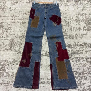 Könnte beinhalten: Blaue Jeans mit Patchwork-Details. Die Jeans haben bordeauxfarbene und braune Patches mit gestickten Blumenmustern. Die Jeans sind am unteren Ende ausgestellt und hängen an einem Holzbügel.