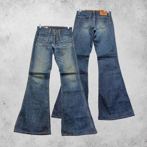 Könnte beinhalten: Zwei Paar blaue Schlaghosen aus Denim. Das vordere Paar hat einen Knopfverschluss und zwei aufgesetzte Vordertaschen. Das hintere Paar zeigt ein klassisches Fünf-Taschen-Design mit einem beigen Lederaufnäher und einem kleinen roten Etikett.