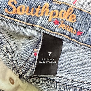 May include: Close-up of blue denim jeans with the brand name "Southpole Jeans" embroidered in orange thread. The jeans have a size 7 label and are made in China. The denim has a faded wash.