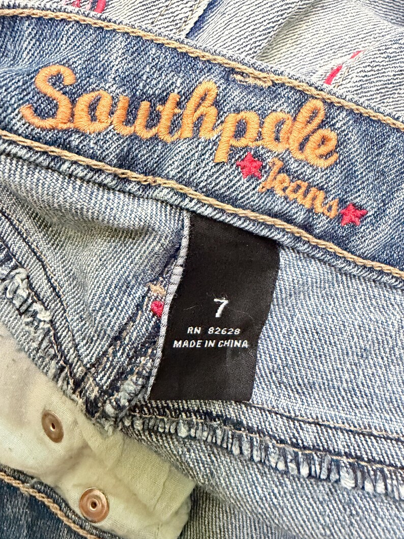 May include: Close-up of blue denim jeans with the brand name "Southpole Jeans" embroidered in orange thread. The jeans have a size 7 label and are made in China. The denim has a faded wash.