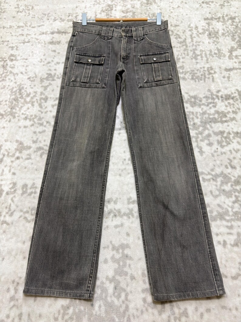 May include: A pair of gray denim pants with two front pockets and a button and zipper closure. The pants are hanging on a hanger against a light-colored background. The pants have a straight leg design.