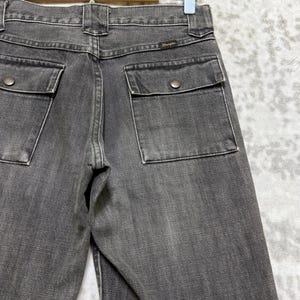 May include: Close-up of the back of a pair of gray denim jeans. The jeans have two square back pockets with button closures and a small Wrangler label. The jeans are hanging on a hanger.