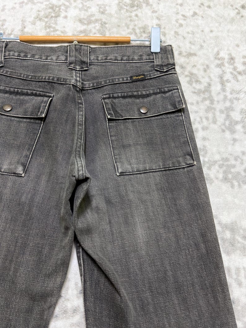 May include: Close-up of the back of a pair of gray denim jeans. The jeans have two square back pockets with button closures and a small Wrangler label. The jeans are hanging on a hanger.