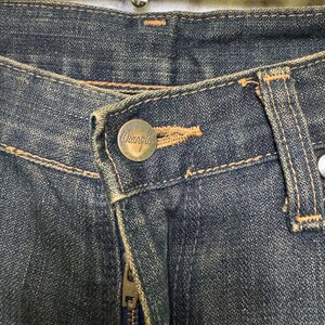 May include: Close-up of a pair of blue denim jeans. The jeans feature a button with the brand name "Wrangler" and a metal zipper. The denim has a worn, faded appearance with visible stitching.