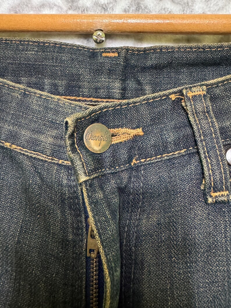 May include: Close-up of a pair of blue denim jeans. The jeans feature a button with the brand name "Wrangler" and a metal zipper. The denim has a worn, faded appearance with visible stitching.