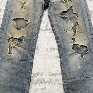 Puede incluir: Vaqueros de mezclilla azul claro con detalles desgastados, con grandes rasgaduras y parches. Los vaqueros tienen un lavado descolorido y un desgaste visible, mostrando una est&eacute;tica vintage. El dise&ntilde;o incluye un estilo cl&aacute;sico de cinco bolsillos y cierre de bot&oacute;n y cremallera.