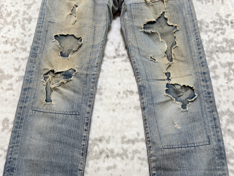 Puede incluir: Vaqueros de mezclilla azul claro con detalles desgastados, con grandes rasgaduras y parches. Los vaqueros tienen un lavado descolorido y un desgaste visible, mostrando una est&eacute;tica vintage. El dise&ntilde;o incluye un estilo cl&aacute;sico de cinco bolsillos y cierre de bot&oacute;n y cremallera.