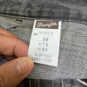May include: Close-up of a pair of gray Wrangler jeans. The tag reads "Wrangler" and includes size information: W5502, 28, W71, H88, WCM-059, and F683-1310. The jeans are made of a textured denim fabric.