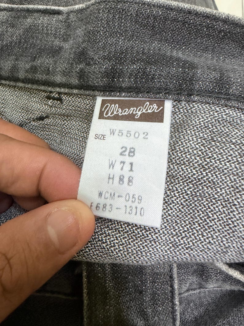 May include: Close-up of a pair of gray Wrangler jeans. The tag reads "Wrangler" and includes size information: W5502, 28, W71, H88, WCM-059, and F683-1310. The jeans are made of a textured denim fabric.