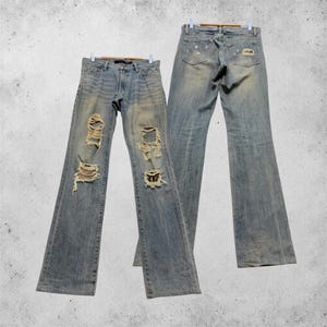 May include: A pair of light-wash blue denim jeans featuring a distressed finish with multiple frayed rips on the front legs and a subtle faded effect. The back view shows two rear pockets with additional distressed detailing and frayed patches.