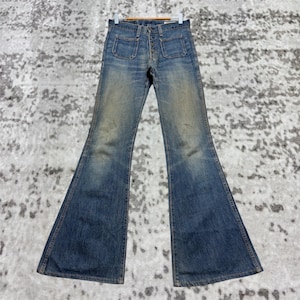 Könnte beinhalten: Vintage blaue Jeans mit Schlaghose aus Denim mit Knopfleiste und zwei Vordertaschen. Die Jeans sind verblichen und weisen Gebrauchsspuren auf. Die Jeans hängen an einem Holzkleiderbügel.