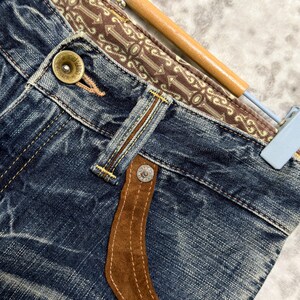 May include: Close-up of blue denim jeans with a brown suede pocket detail and decorative stitching. The waistband features a patterned brown fabric. A gold button and silver rivets are visible.