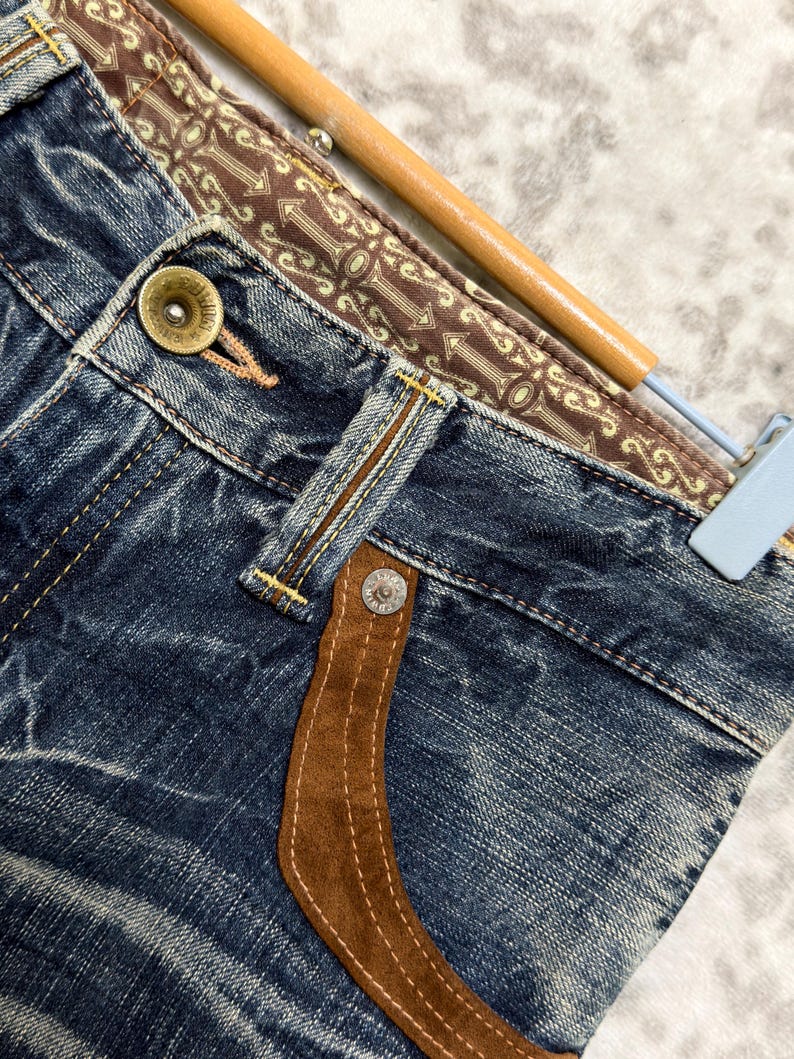 May include: Close-up of blue denim jeans with a brown suede pocket detail and decorative stitching. The waistband features a patterned brown fabric. A gold button and silver rivets are visible.