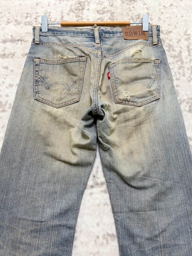 Puede incluir: Vaqueros de mezclilla azul descolorido con detalles desgastados, incluyendo rasgaduras y marcas de uso. Los bolsillos traseros presentan un dise&ntilde;o cl&aacute;sico, y una etiqueta de cuero con la marca "EDWIN" es visible. Los vaqueros est&aacute;n colgados en una percha de madera.