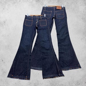 Könnte beinhalten: Zwei Paar dunkelblaue Jeans aus Indigo-Denim mit ausgestelltem Beinverlauf. Das vordere Paar hat einen Knopfverschluss und zwei aufgesetzte Taschen. Das hintere Paar zeigt zwei Gesäßtaschen und einen braunen Lederaufnäher am Bund.