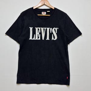 Pode incluir: T-shirt preto com o texto branco "LEVI'S" no peito. A camisa tem mangas curtas e uma pequena etiqueta vermelha na parte inferior esquerda. A camisa está pendurada em um cabide de madeira.