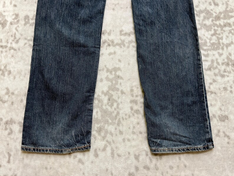 May include: Close-up of a pair of dark blue denim jeans. The jeans have a straight leg cut and are made of a textured denim fabric. The hem of the jeans is visible, showing the stitching detail.