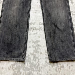 May include: A close-up shot of a pair of gray denim jeans. The jeans have a straight leg cut and are made of a faded gray denim material. The stitching is visible along the seams and pockets. The jeans are laid flat.