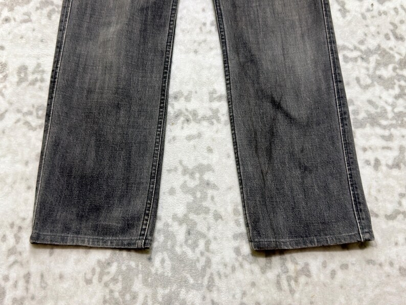 May include: A close-up shot of a pair of gray denim jeans. The jeans have a straight leg cut and are made of a faded gray denim material. The stitching is visible along the seams and pockets. The jeans are laid flat.