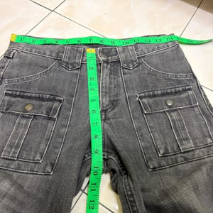 May include: Close-up of gray denim pants with two front pockets and button closures. A green measuring tape shows the waist size. The pants are made of a durable fabric and are suitable for casual wear.