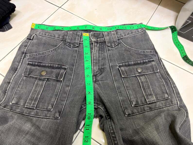 May include: Close-up of gray denim pants with two front pockets and button closures. A green measuring tape shows the waist size. The pants are made of a durable fabric and are suitable for casual wear.