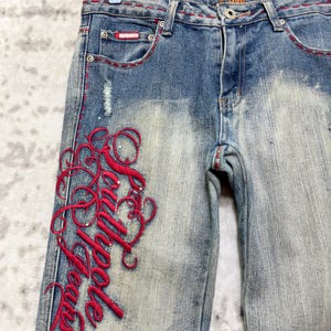 May include: Light blue denim jeans with red stitching and embroidered script on the left leg. The jeans have a distressed wash and feature a small red patch on the front pocket.