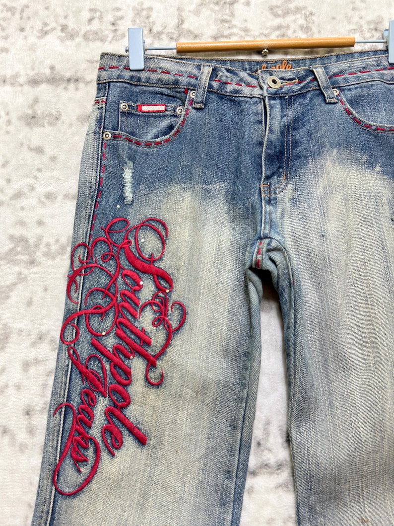 May include: Light blue denim jeans with red stitching and embroidered script on the left leg. The jeans have a distressed wash and feature a small red patch on the front pocket.