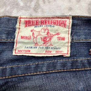 May include: Close-up of a blue denim jeans label. The label is rectangular with the text "TRUE RELIGION BRAND JEANS" in red. A red graphic of a Buddha playing a guitar is in the center. The text "WORLD TOUR" is also visible.