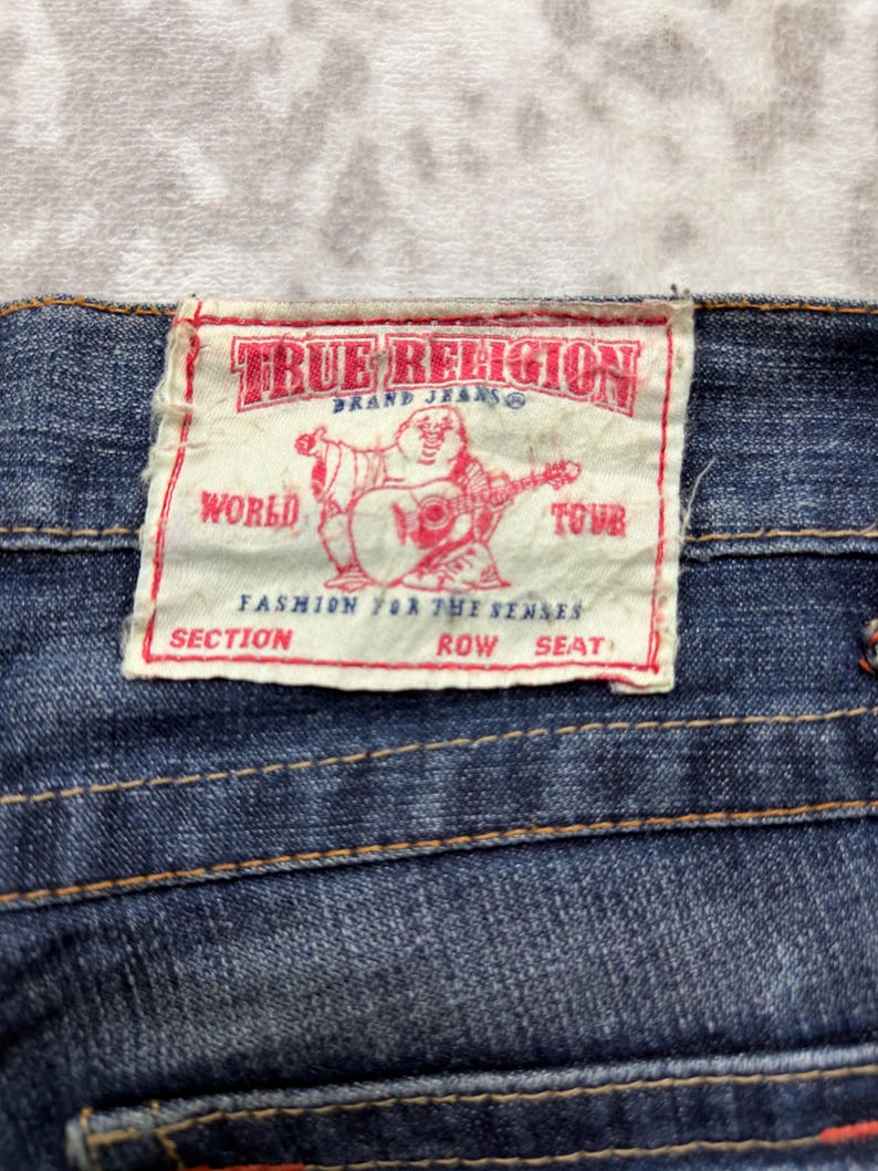 May include: Close-up of a blue denim jeans label. The label is rectangular with the text "TRUE RELIGION BRAND JEANS" in red. A red graphic of a Buddha playing a guitar is in the center. The text "WORLD TOUR" is also visible.
