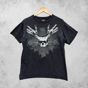 Könnte beinhalten: Schwarzes T-Shirt mit einem Grafikdesign, das einen Totenkopf, stilisierte Hände und dekorative Elemente zeigt. Der Text "Legend Killer" ist sichtbar. Das T-Shirt hängt an einem Holzbügel.