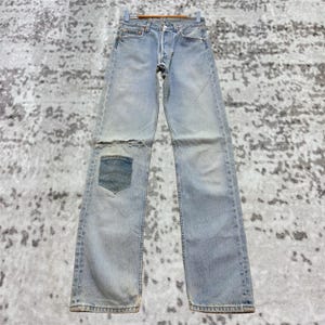 Könnte beinhalten: Hellblaue Jeans mit Used-Look und geflicktem Knie. Die Jeans hängen an einem Holzkleiderbügel. Der Denim hat ein verblasstes Aussehen mit sichtbaren Gebrauchsspuren.