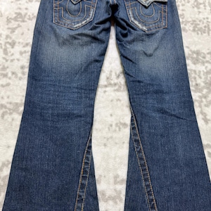 May include: Blue denim jeans with a flared leg design. The jeans feature classic five-pocket styling, with decorative stitching on the back pockets. The denim has a faded wash and distressed details, giving them a worn look.