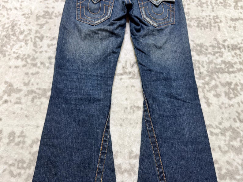 May include: Blue denim jeans with a flared leg design. The jeans feature classic five-pocket styling, with decorative stitching on the back pockets. The denim has a faded wash and distressed details, giving them a worn look.