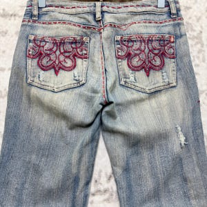 May include: Light blue denim jeans with red decorative stitching and embroidered pocket details. The jeans have a distressed look with a faded wash and a small tear on the right leg.