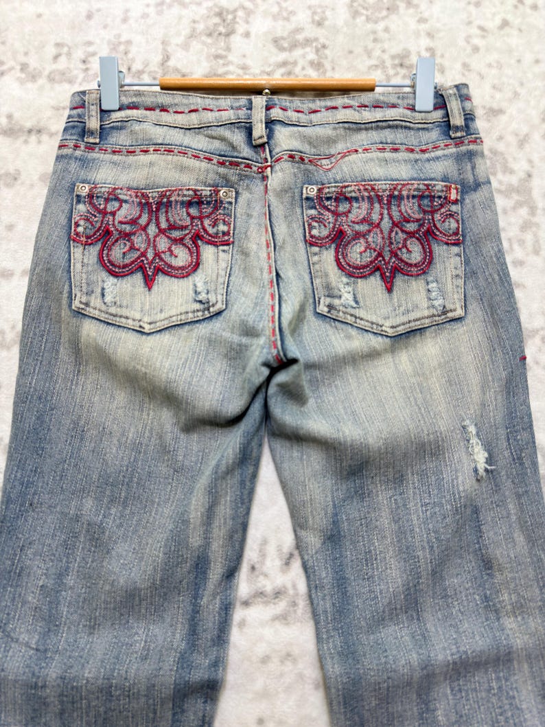 May include: Light blue denim jeans with red decorative stitching and embroidered pocket details. The jeans have a distressed look with a faded wash and a small tear on the right leg.