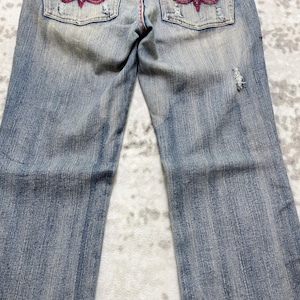 May include: Faded blue denim jeans with decorative red stitching and embellishments on the back pockets. The jeans have a distressed look with a small tear on the right leg.