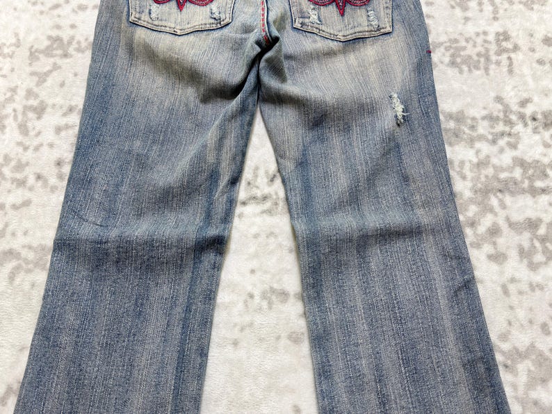 May include: Faded blue denim jeans with decorative red stitching and embellishments on the back pockets. The jeans have a distressed look with a small tear on the right leg.