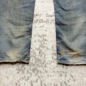 Puede incluir: Primer plano de la parte inferior de unos vaqueros azules descoloridos. La mezclilla muestra desgaste, con parches m&aacute;s claros y zonas desgastadas. El dobladillo de los vaqueros est&aacute; ligeramente deshilachado.