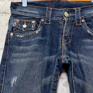May include: Close-up of dark blue denim jeans with orange stitching and silver hardware. The jeans have a classic five-pocket design, with a button and zipper closure. The jeans are distressed, with some fading and wear.