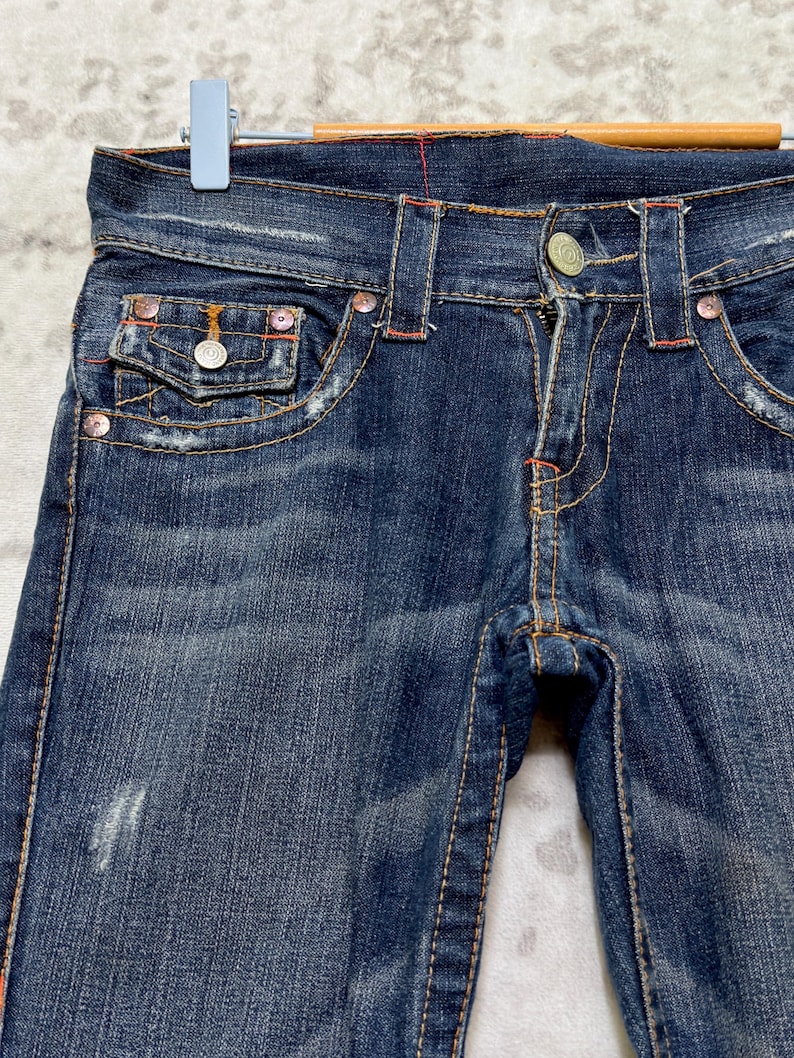 May include: Close-up of dark blue denim jeans with orange stitching and silver hardware. The jeans have a classic five-pocket design, with a button and zipper closure. The jeans are distressed, with some fading and wear.