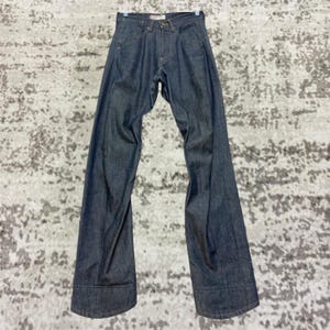 Puede incluir: Vaqueros de mezclilla de lavado oscuro con pernera acampanada. Los vaqueros tienen un diseño clásico de cinco bolsillos, cierre de botón y cremallera y trabillas para el cinturón. La mezclilla es de color índigo oscuro y los vaqueros se muestran en una percha.