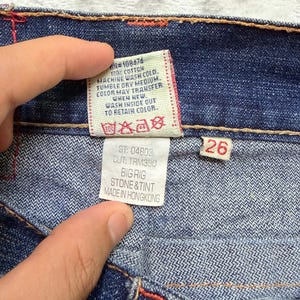 May include: Close-up of blue denim jeans with red stitching. A white tag displays washing instructions and the text "BIG RIG STONE & TINT MADE IN HONGKONG". A red tag shows the size "26".