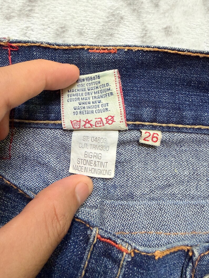 May include: Close-up of blue denim jeans with red stitching. A white tag displays washing instructions and the text "BIG RIG STONE & TINT MADE IN HONGKONG". A red tag shows the size "26".