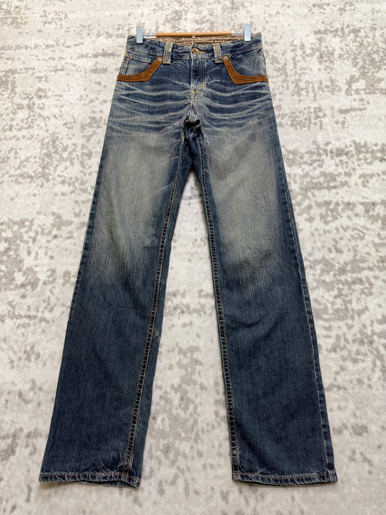 May include: Blue denim jeans with a faded wash and brown suede pocket accents. The jeans have a classic five-pocket design and a straight leg fit. The jeans are hanging on a wooden hanger.