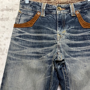 May include: Close-up of blue denim jeans with brown suede pocket accents. The jeans have a faded wash and a decorative patterned waistband. The jeans are hanging on a wooden hanger.