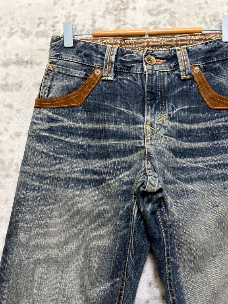 May include: Close-up of blue denim jeans with brown suede pocket accents. The jeans have a faded wash and a decorative patterned waistband. The jeans are hanging on a wooden hanger.