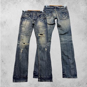 Op de afbeelding: Twee paar blauwe spijkerbroeken van denim met een versleten afwerking, gerafelde gaten en waspatronen. De broeken hebben een klassiek ontwerp met vijf zakken, metalen knoopsluitingen en zichtbare stiksels op de achterzakken en de broekspijpen.